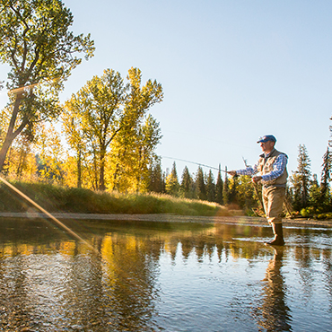 A man standing in shallow water holding a fishing rod, and trees in the background.