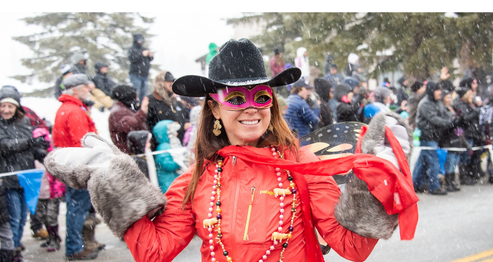 A woman in a red jacket with giant mittens smiles and walks through a snowy parade.
