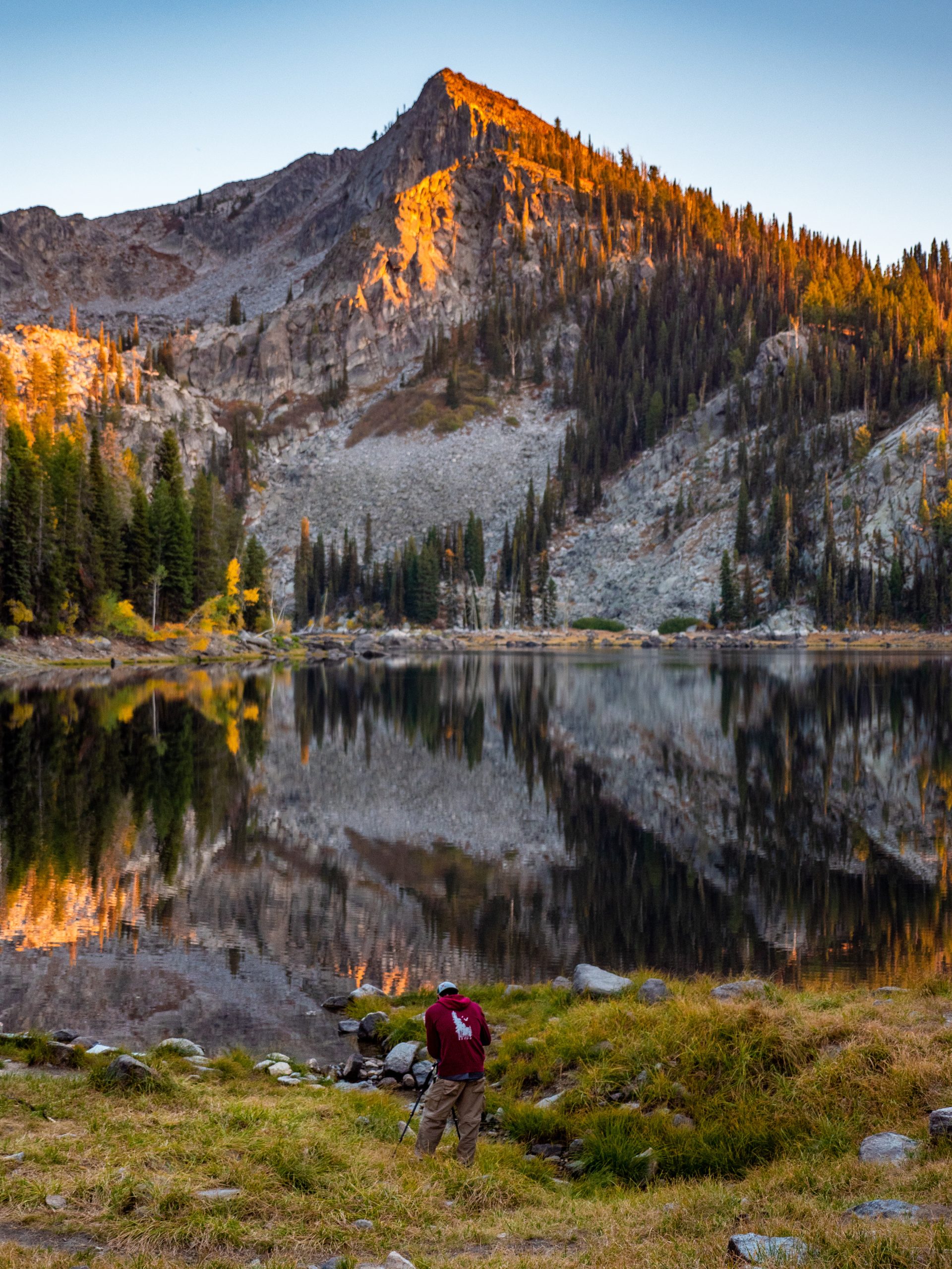 A person standing in front of lake beside a tree-covered mountain.