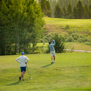 Two men on a golf course surrounded by tall trees.