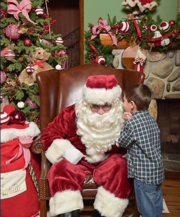 A boy in a checkered shirt whispers into Santa's ear while he sits in a big chair. 