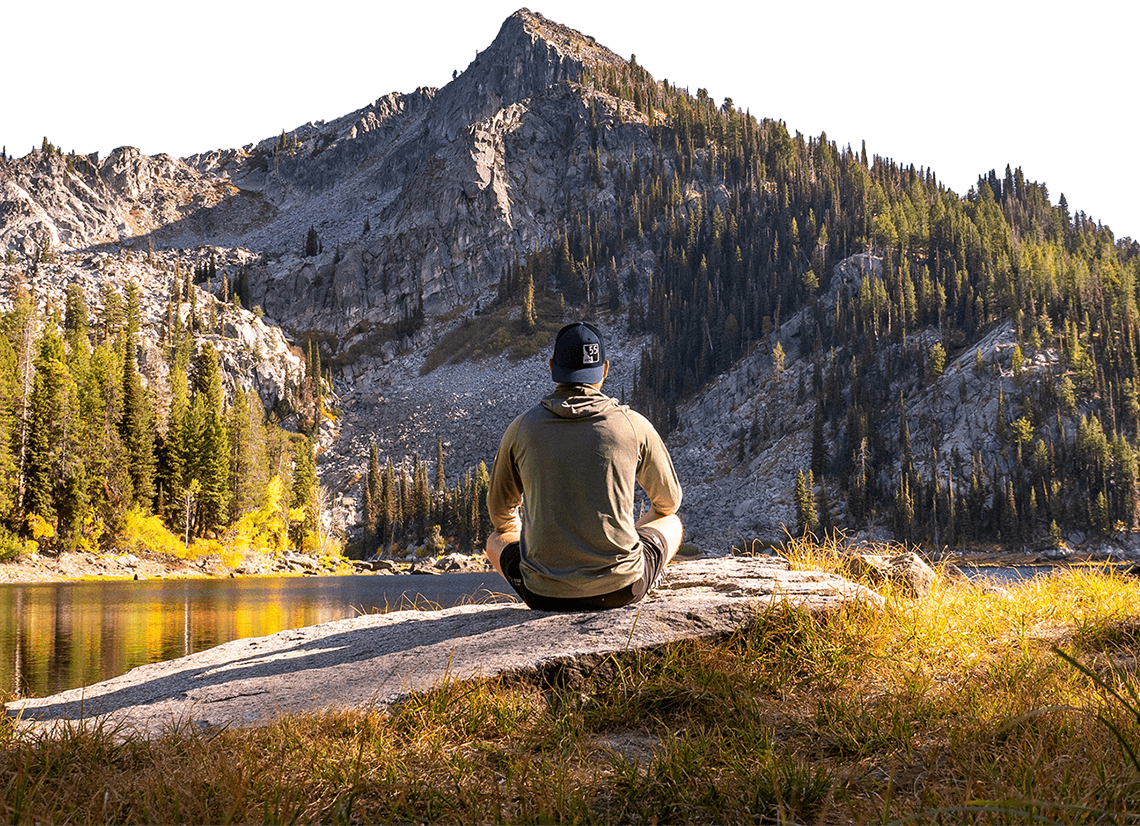 Person sitting on a rock by a lake with tall trees and a rugged mountain in the background.