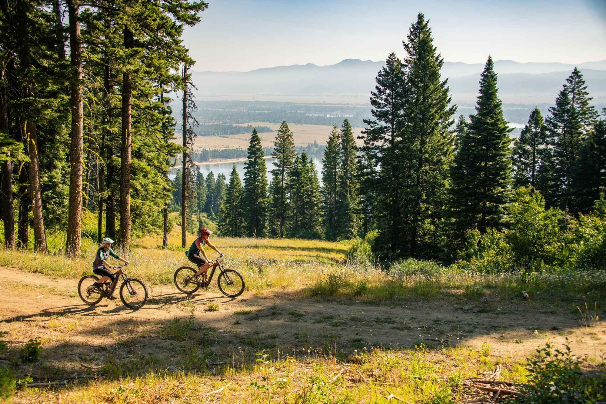 Two people mountain biking on a trail at Tamarack surrounded by tall grass and a forest of trees, and body of water and mountains in the distance.