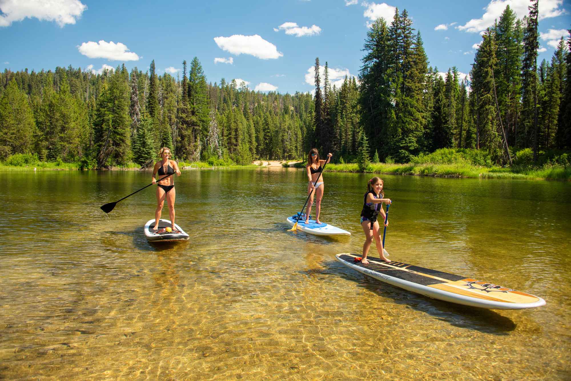 Two women and a child stand up paddleboarding on a lake surrounded by a thick forest of trees.