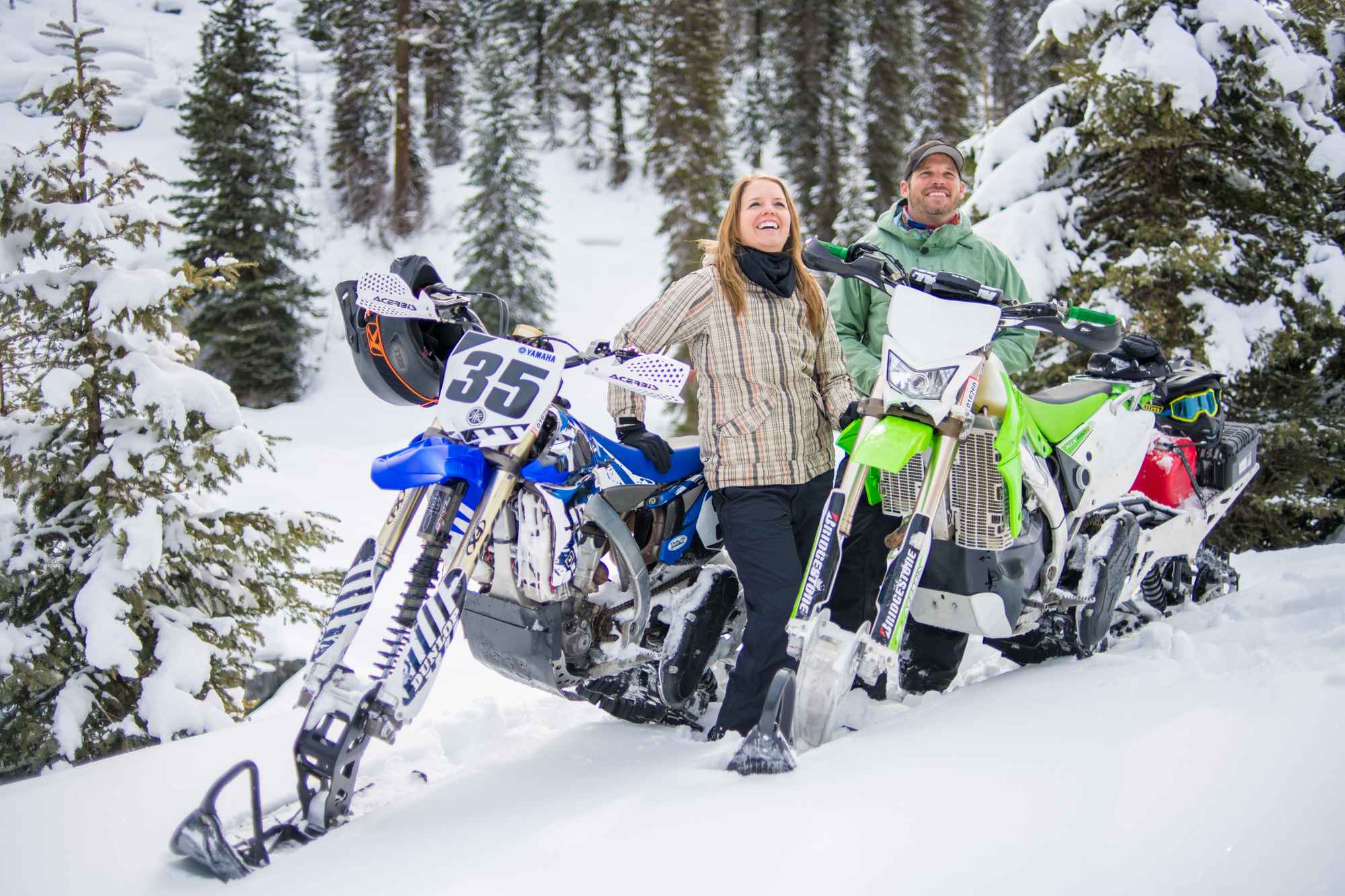 Two people standing beside two snowmobiles in a field of snow backed by a snow-covered forest of trees.