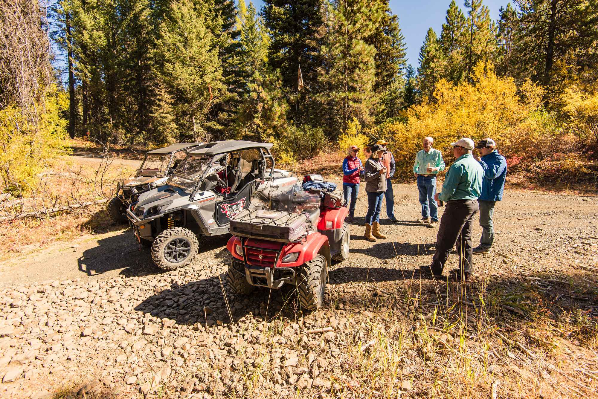 A group of people standing on a dirt road behind an ATV and two UTVs, surrounded by a forest of trees.